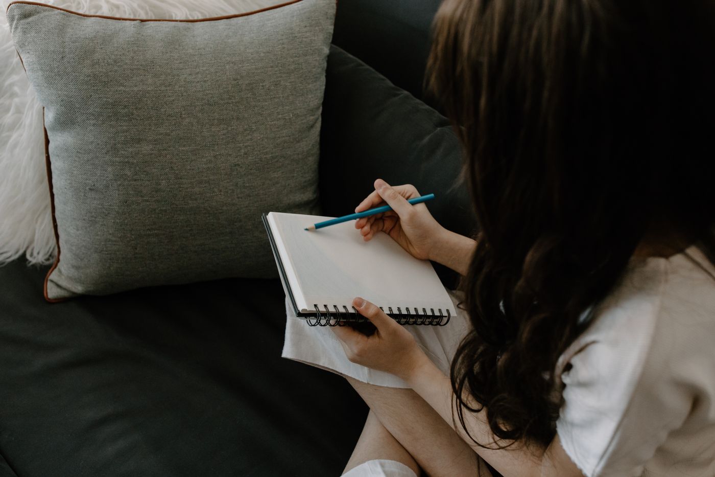 Pessoa escrevendo no caderno o que precisa, sentada no sofá de casa.